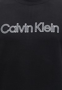 Tissu noir avec texte brodé blanc affichant le nom de la marque "Calvin Klein" en lettres claires et grasses au centre.
