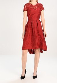 Robe rouge à manches courtes, haut en dentelle, tissu texturé, jupe plissée et ourlet asymétrique, associée à des talons noirs.