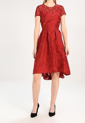 Red dress with short sleeves, lace top, textured fabric, pleated skirt, and high-low hem, paired with black heels.