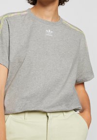 Grå bomullströja med korta ärmar, med flerfärgade randiga detaljer på axlarna och en vit Adidas-logotyp på bröstet.