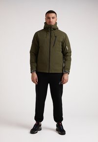 Oliven grøn isoleret jakke med hætte, frontlynlås og sorte accenter. Bæres med sorte sweatpants og sorte sneakers.