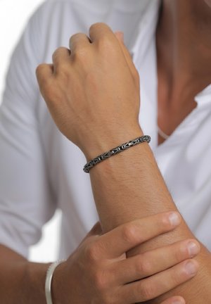 Zilveren armband met een ingewikkeld geweven ontwerp, een gestructureerd oppervlak en een gepolijste afwerking, gedragen aan een licht gebruinde pols.