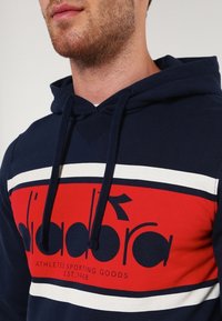 Haut à capuche bleu marine avec des rayures horizontales rouges et blanches, comportant un imprimé logo sur la poitrine et une capuche avec cordon. Matière en coton, coupe décontractée.