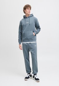 Grå hættetrøje og matchende sweats, lavet af blødt stof, med en frontlomme og logo detalje. Sammen med sorte og hvide sneakers.