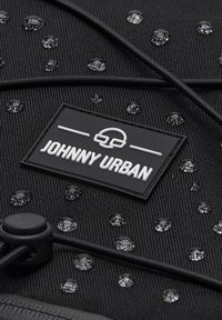Fekete textúrájú anyag vízcseppekkel, elasztikus zsinór, kapcsoló, és egy téglalap alakú "Johnny Urban" logó címke fehér betűkkel.