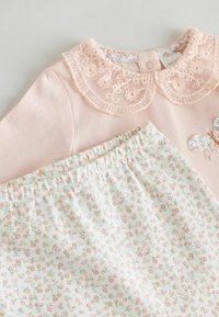 Top rosa claro con cuello de encaje y aplicación floral, combinado con pantalones blancos con un pequeño estampado floral y cintura elástica.