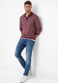 Polo a manica lunga color marrone con colletto rosso, dettagli con tasca, abbinato a jeans blu e sneaker bianche. Tessuto liscio e vestibilità comoda.