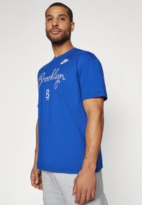 Mėlynas medvilninis marškinėlis su užrašu „Brooklyn“ baltu šriftu ir Nike logotipu ant krūtinės. Trumpomis rankovėmis, standartinio kirpimo, minimaliai tekstūruotas. Matomos pilkos šortai.