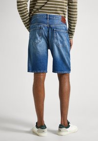 Persona vistiendo pantalones cortos de mezclilla azul de longitud media, una camisa de manga larga a rayas en verde oliva y blanco, y zapatillas blancas con acentos en verde oscuro.