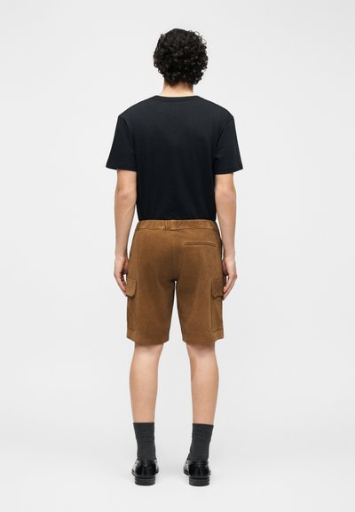 Homme aux cheveux bouclés portant un t-shirt noir, un short cargo marron, des chaussettes foncées et des chaussures noires, debout de dos sur un fond uni.