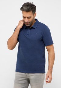 Tamsiai mėlynas polo marškinėliai trumpomis rankovėmis, su trimis mygtukais, subtiliu tekstūru ir maža logotipu ant krūtinės. Derinami su šviesiai pilkomis kelnėmis.