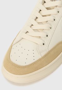 Baskets beiges avec un mélange de cuir lisse et de suède. Le design comprend des perforations sur le bout, une languette en textile et des lacets plats.