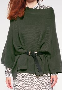 Olivgrön ribbad poncho med ett svart bälte, bärs över en mönstrad klänning. Har breda ärmar och en lös passform. Texturerad tyg.