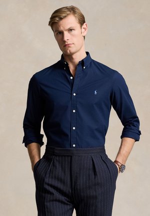 Man wearing navy blue button-up shirt with rolled sleeves and pinstriped trousers, hands in pockets, beige background.