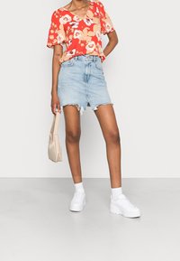 Top rojo con estampado floral y mangas cortas, combinado con una minifalda de mezclilla clara con dobladillo deshilachado y una abertura frontal. Zapatillas blancas y un bolso beige.