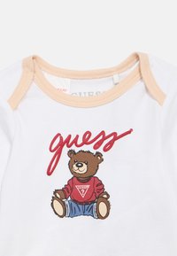 Λευκό πουκάμισο με κοντά μανίκια και μπεζ περίγραμμα, με σχέδιο ενός καρτούν αρκούδου που φοράει κόκκινο πουλόβερ και μπλε τζιν, με το λογότυπο "guess" τυπωμένο πάνω.