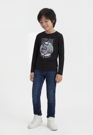 Uśmiechnięty chłopiec stoi z rękami opartymi wzdłuż ciała, ubrany w czarną bluzę z długim rękawem z grafiką węża i napisem "EXPLORE", niebieskie dżinsy i białe trampki.