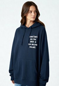 Sweat à capuche oversize bleu marine en molleton, avec une poche avant et un texte blanc indiquant « Parfois, tout ce dont vous avez besoin, c’est de 100 millions de dollars. »