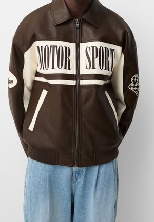 Persona con chaqueta de cuero marrón y crema con texto "MOTOR SPORT" y parche de bandera a cuadros en las mangas, combinada con vaqueros azul claro.