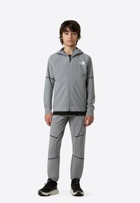 Grå treningsjakke og -bukser med svarte detaljer; lettvekt jakke med glidelås og hette, bukser med mansjetter, reflekterende logo på brystet.