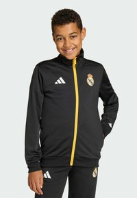 Musta vetoketjullinen urheilutakki, jossa on keltainen vetoketju ja Real Madridin logo, valkoisilla Adidas-raidoilla hihoissa ja kalvosimissa, valmistettu sileästä kankaasta.