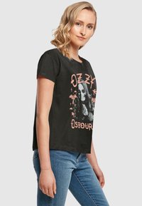 Sort bomulds t-shirt med korte ærmer, med et grafisk print af en person med solbriller og teksten "OZZY OSBOURNE" i orange.
