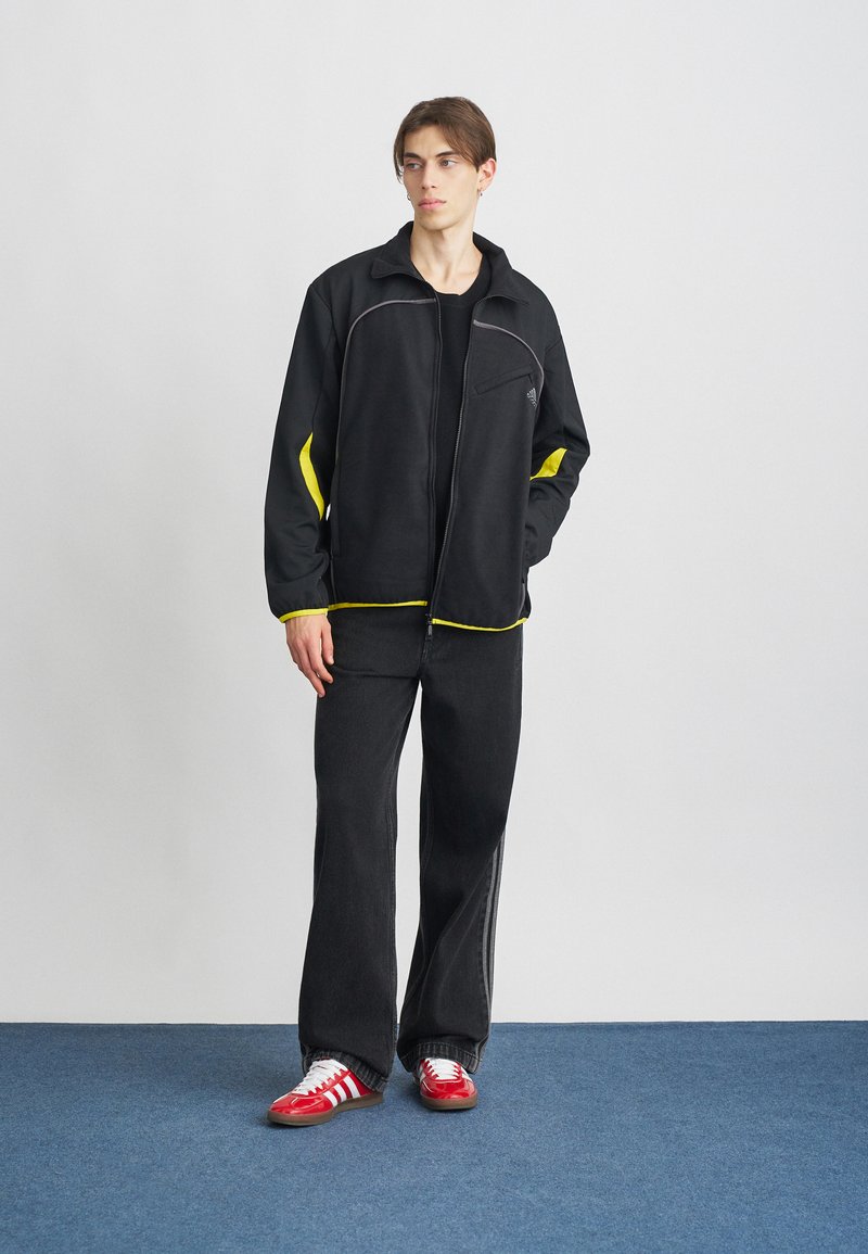 Schwarze Zip-Jacke mit gelben Akzenten, kombiniert mit schwarzen weit geschnittenen Hosen und roten Turnschuhen mit weißen Streifen. Glatte Textur und lässiges Design.