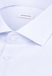 Gros plan sur le col d'un polo blanc texturé avec deux boutons blancs et une étiquette de marque "seidensticker" à l'intérieur de l'encolure.