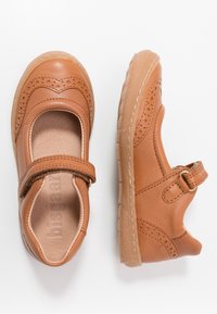 Zapatos de cuero marrón con punta redondeada, acentos perforados y una correa de Velcro. Suela de goma flexible con un patrón texturizado.