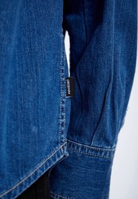 Denim jacket in dark blue with a textured surface, featuring a black label reading "DRDENIM" near the seam on the sleeve.