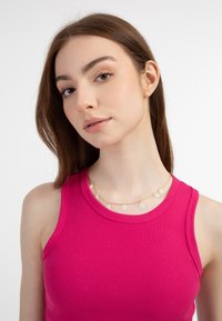 Collana d'oro con charm a margherita bianca, indossata sopra una canotta rosa a costine. Gli accenti a forma di margherita sono equidistanti lungo una delicata catenina.