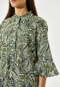Vihreä ja kerma valkoinen kuviollinen blouse korkealla kauluksella, nappilukituksella ja levenevillä hihoilla, jossa on kasviaiheisia kuvioita ja monimutkaista yksityiskohtaa.