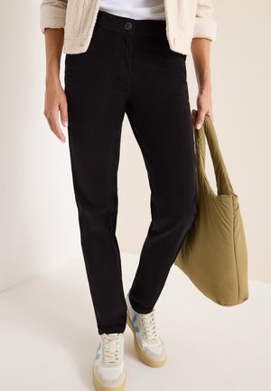 Pantalones de pana negros con cierre de botón, corte recto y textura sutil; combinados con un bolso acolchado verde y zapatillas casuales.