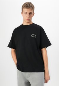 Ung mand med kort blond hår iført en sort oversized T-shirt med lille hvid tekst på brystet og lysegrå bukser, stående imod en hvid væg.