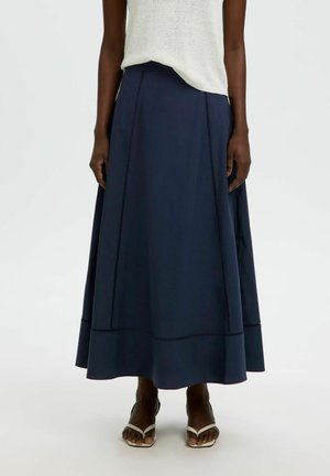Femme porte une jupe midi évasée bleu marine avec des détails de couture verticale, associée à des sandales blanches à brides et un top léger sans manches.
