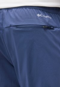 Σορτς Columbia σε ναυτικό μπλε με ελαστική μέση και πίσω τσέπη με φερμουάρ, που τα φορά ένα άτομο ορατό από τη μέση και κάτω.