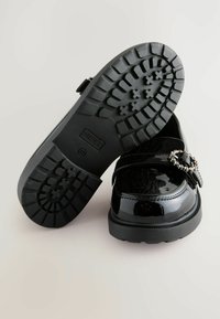 Mocasiini din piele patent neagră, cu o talpă din cauciuc texturat, cataramă decorativă argintie și un profil jos pentru confort și stabilitate.