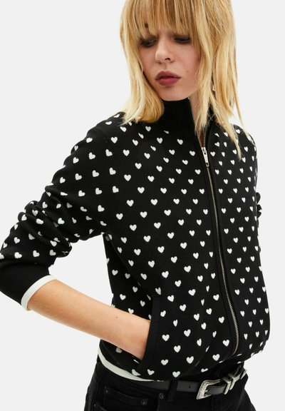 Pull noir zippé avec un motif de cœurs blancs, des poignets côtelés et un col montant, fabriqué en maille douce.