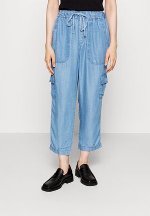 Femme portant un pantalon cargo court bleu clair avec une taille à cordon de serrage et des mocassins en cuir noir, debout devant un fond uni.
