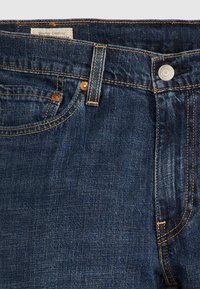 Jeans de mezclilla azul oscuro que muestran la cinturilla, el botón, la trabilla del cinturón, el bolsillo delantero con remaches y la costura de la etiqueta visible.