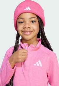 Růžový pulovr s čtvrt zipem, vyrobený z texturované látky, s bílým logem Adidas na prsou a odpovídající růžovou pletenou čepicí.