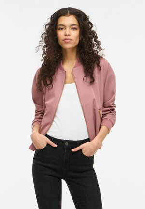 Femme aux cheveux bouclés portant une veste zippée rose poudré sur un haut blanc et un jean noir, debout les mains dans les poches sur un fond blanc.