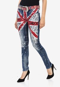 Blaue Jeans aus Denim mit einem Union Jack-Design in Rot und Weiß, verziert mit Farbklecksen. Enthält einen mit Nieten besetzten silbernen Gürtel und einen figurschmeichelnden Schnitt.