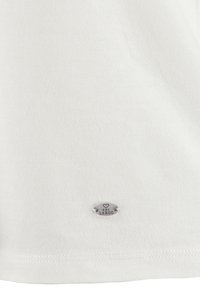 Camiseta de algodón blanca con una textura suave, que presenta una pequeña insignia plateada con el texto "Key Largo" cerca del dobladillo. Diseño simple, sin patrones.