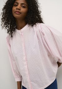 Light pink blouse with textured polka dots, button-down front, and puff sleeves. Soft fabric, gathered detailing at shoulders, and rounded hem.