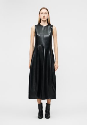 Γυναίκα με ίσια μαλλιά που φοράει ένα μαύρο μίντι φορεμα από ψεύτικο δέρμα χωρίς μανίκια και μαύρα μποτάκια μέχρι τον αστράγαλο, στεκόμενη μπροστά σε απλό λευκό φόντο.