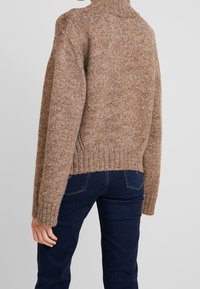 Pull en tricot marron avec un col montant, présentant un tissage texturé et des poignets côtelés, associé à un jean bleu foncé de style ajusté.