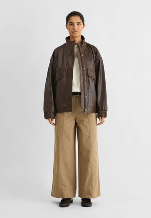 Un mannequin se tient devant un fond de studio uni, vêtu d’une veste en cuir marron foncé, d’un haut couleur crème, d’un pantalon beige ample et de chaussures foncées.
