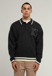 Černý pletený pulovr s bílým pruhovaným límcem a manžetami. Na levé části hrudi je vyšitý dopis 'K' se hvězdami.