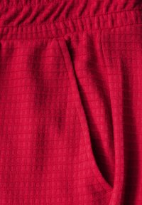 Rode textuur stof met een ruitpatroon, voorzien van een elastische tailleband en een zijzak. Het materiaal lijkt zacht en iets pluizig.